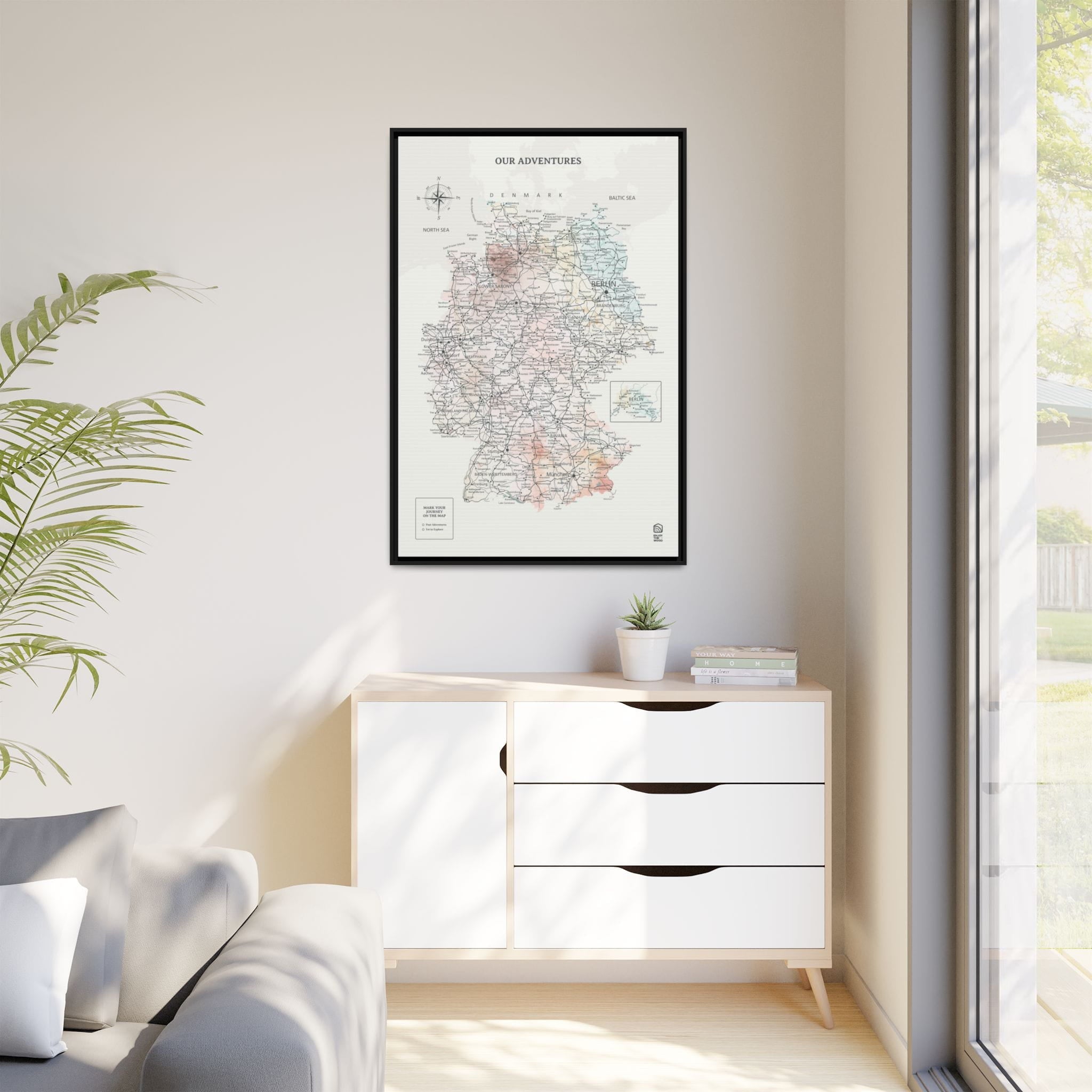 Framed map on a wall above a white cabinet with a plant and books.
