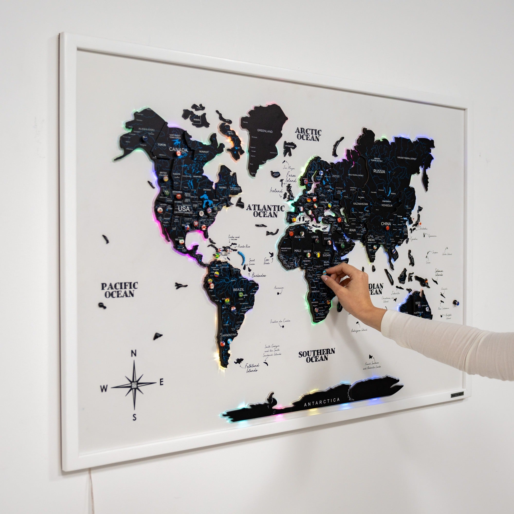 World map with colorful pins on a white wall, hand pointing at a location.